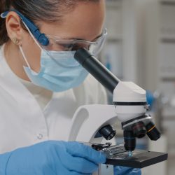Biologist with face mask using microscope in science laboratory, analyzing dna sample and substance with microscopic tool. Woman with protective glasses working with magnifying glass lens